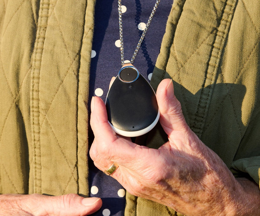 Person holding a black personal alarm with a green jacket and patterned shirt in the background