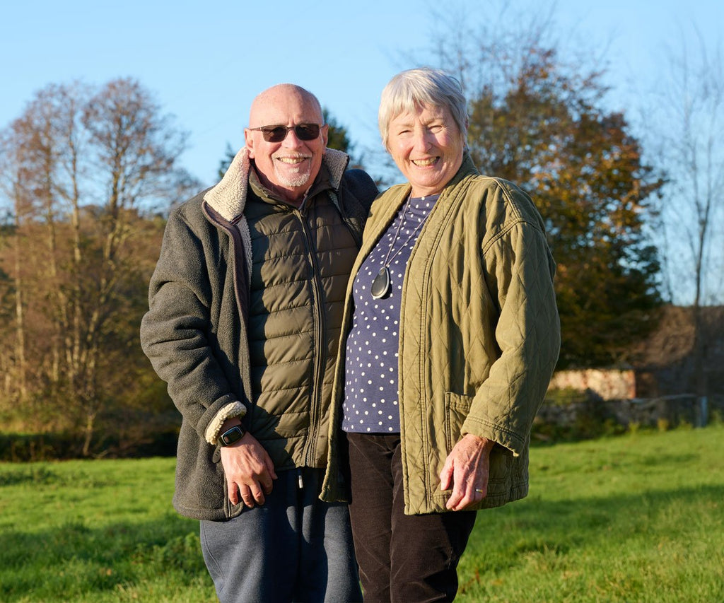 Two people standing in a grassy field with trees in the background, both wearing personal alarms
