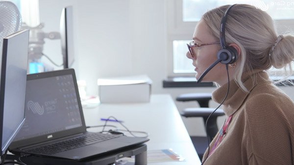 A woman with blonde hair in a bun, wearing glasses and a beige cardigan, is shown in profile wearing a headset and working at a desk with a laptop in a bright office setting.
