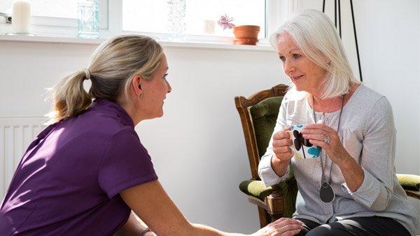 A woman in a purple scrub top sits beside an older woman with white hair sitting in a green armchair, holding sunglasses in a brightly lit room.