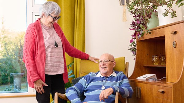 An elderly woman in a pink cardigan and glasses stands beside an elderly man in a blue striped sweater who is sitting in a chair, in a cozy room with a wooden cabinet, plants, and a window.