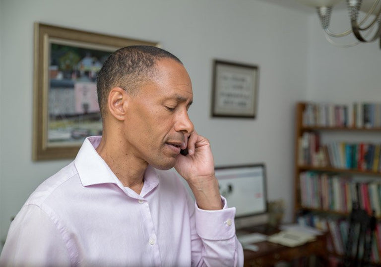 Man talking on a phone in an office setting with a computer and bookshelf in the background.