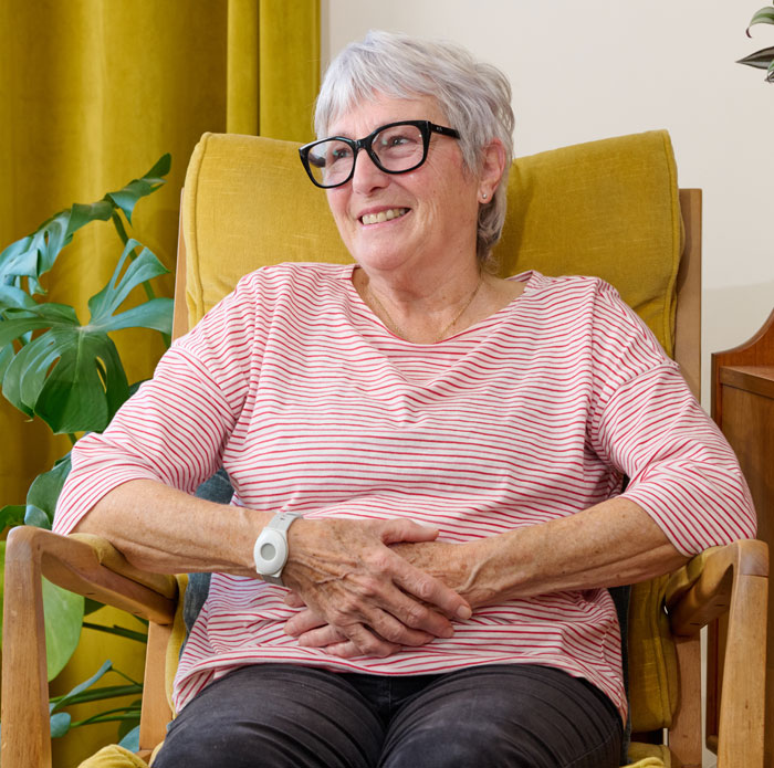 Woman sitting in a yellow armchair wearing a pink striped shirt and glasses, and personal alarm pendant