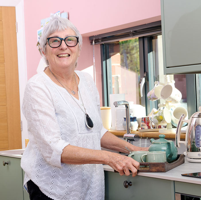 Elderly woman in a kitchen preparing tea, wearing a white shirt and alarm pendant