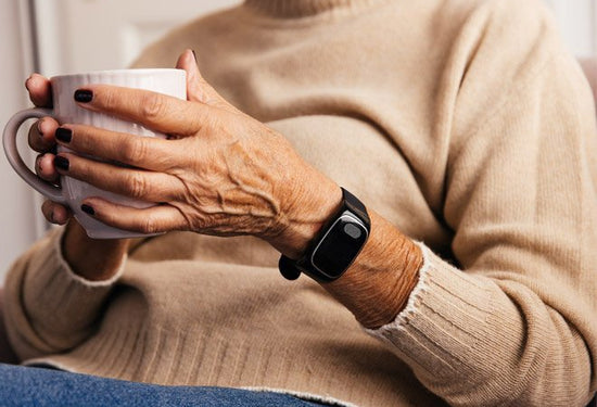 Person wearing a beige sweater holding a white mug with a black dementia tracker on the wrist.
