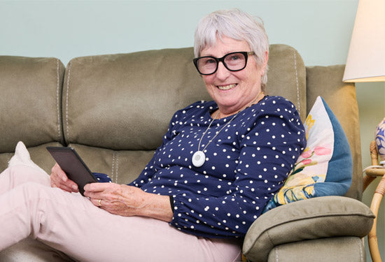 Elderly woman sitting on a couch holding a tablet, wearing glasses and a neck worn alarm pendant