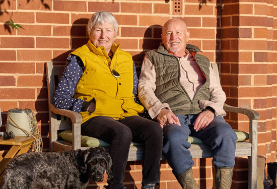 Two elderly people sitting on a bench with a dog, both wearing personal alarms