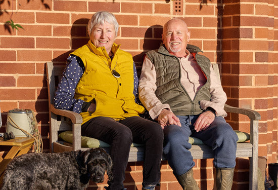 Two elderly people sitting on a bench with a dog, both wearing personal alarms