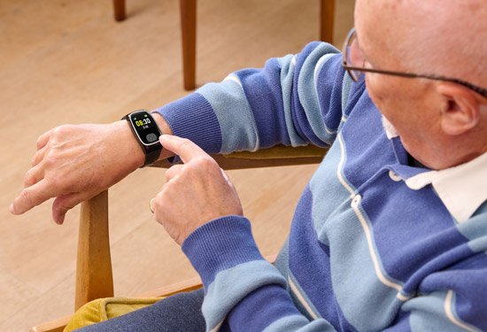 Elderly man wearing a blue sweater using a personal alarm watch on his wrist.