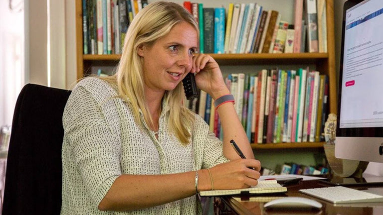 Woman sitting at a desk in an office setting, talking on the phone and taking notes.