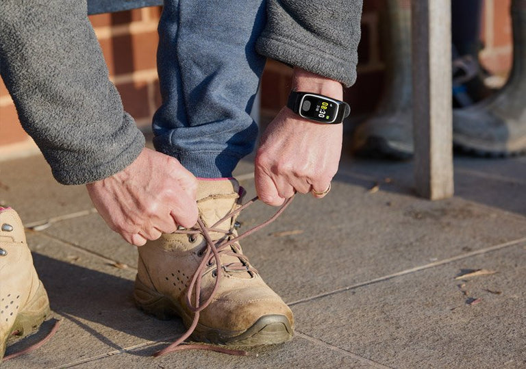 Person tying shoelaces on a brown boot with an alarm smartwatch on the wrist, standing on a sidewalk.