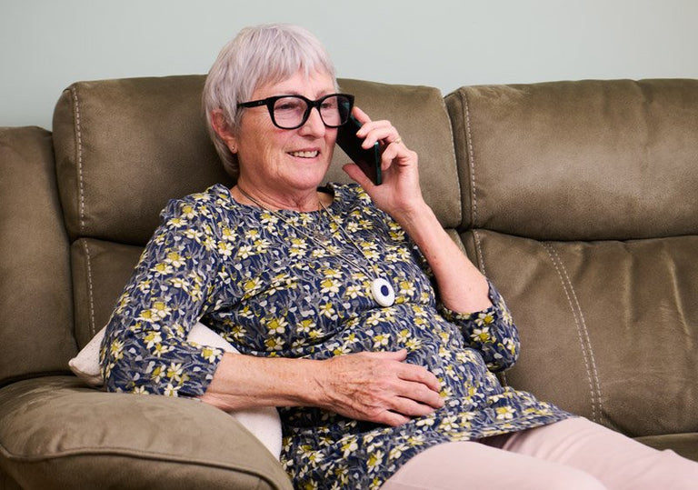 Woman wearing fall alarm sitting on a brown couch talking on a phone