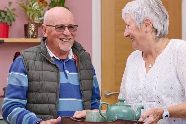 Two elderly people smiling and holding a tray with a teapot indoors.