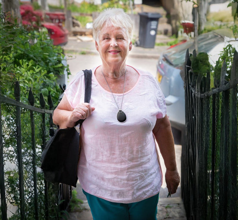 Woman standing outdoors with a black bag, wearing an alarm pendant