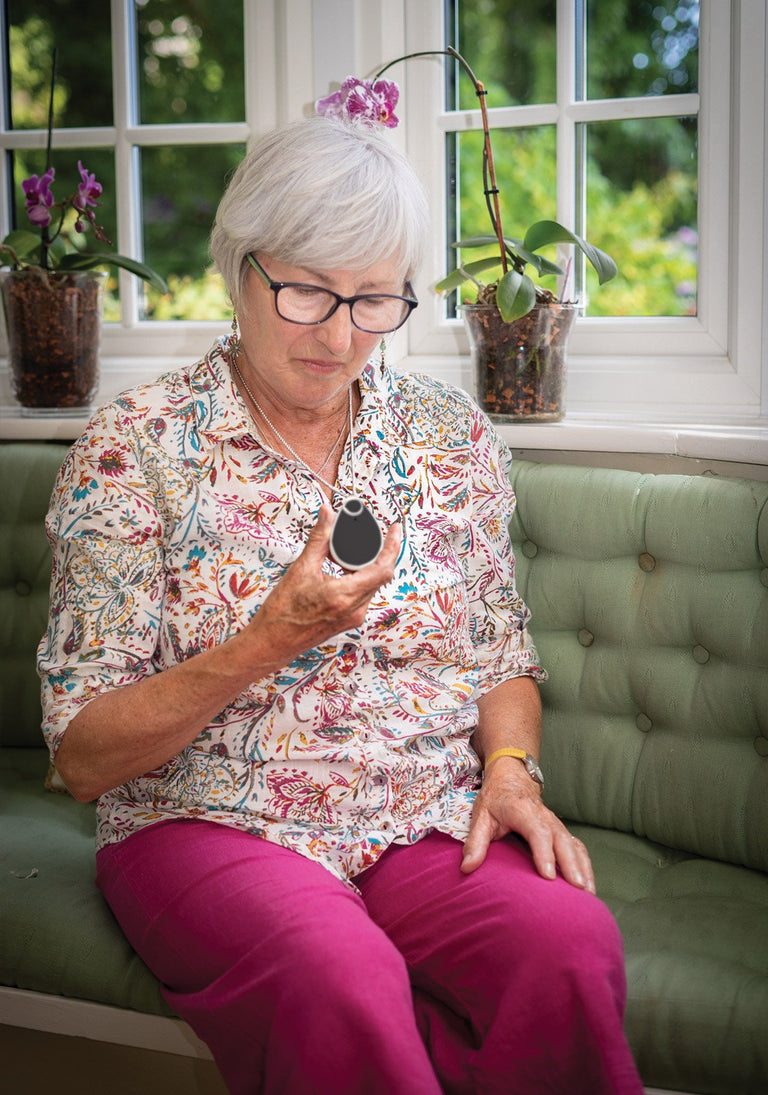 Woman sitting on a green couch holding Taking Care Anywhere pendant, with plants and a window in the background.