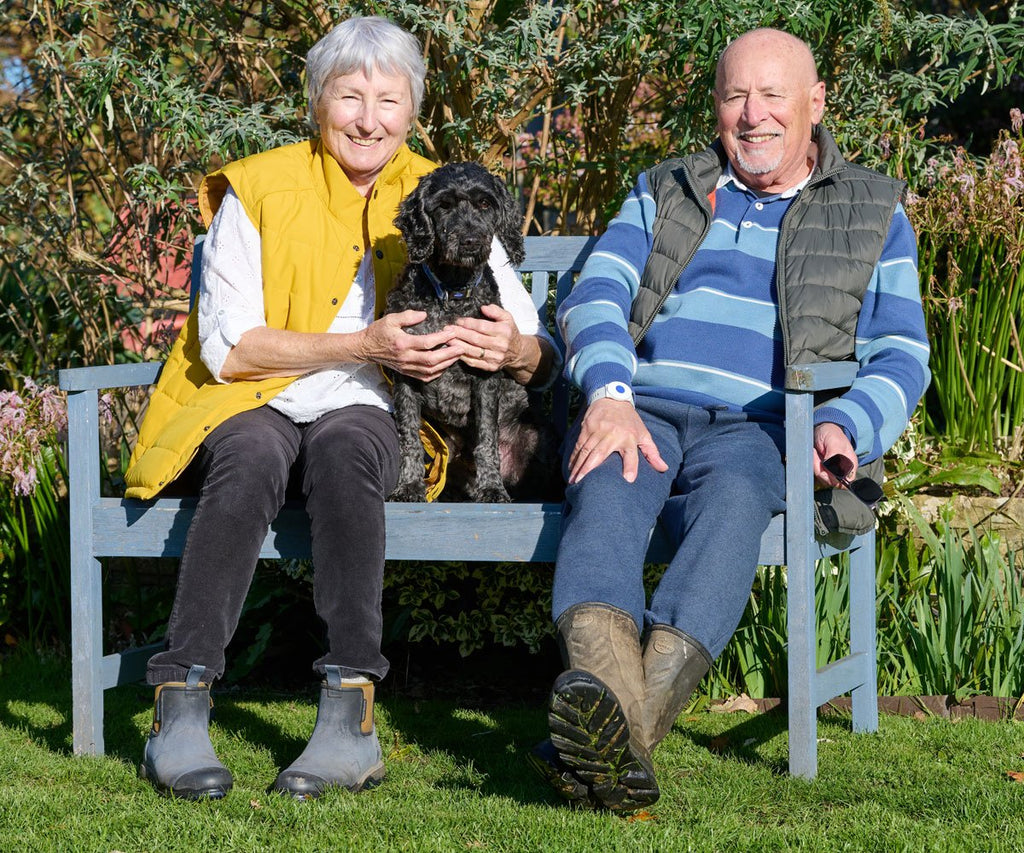 Two elderly people sitting on a bench with a dog in a garden setting