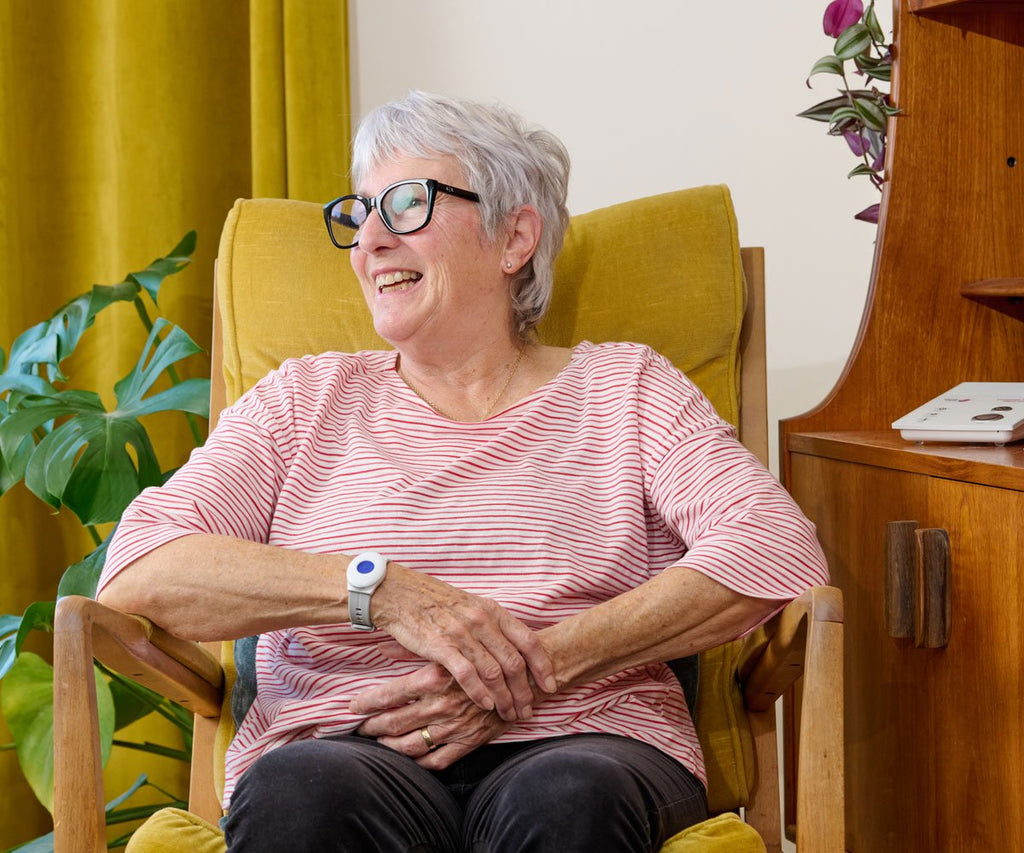 Woman sitting in a yellow armchair wearing glasses and a striped shirt, with a wooden cabinet and plant in the background.