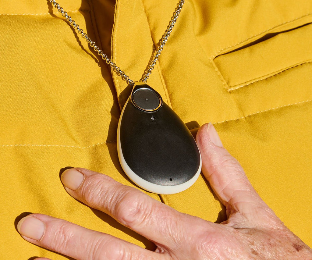 Black pendant alarm on a chain held by a hand against a yellow background
