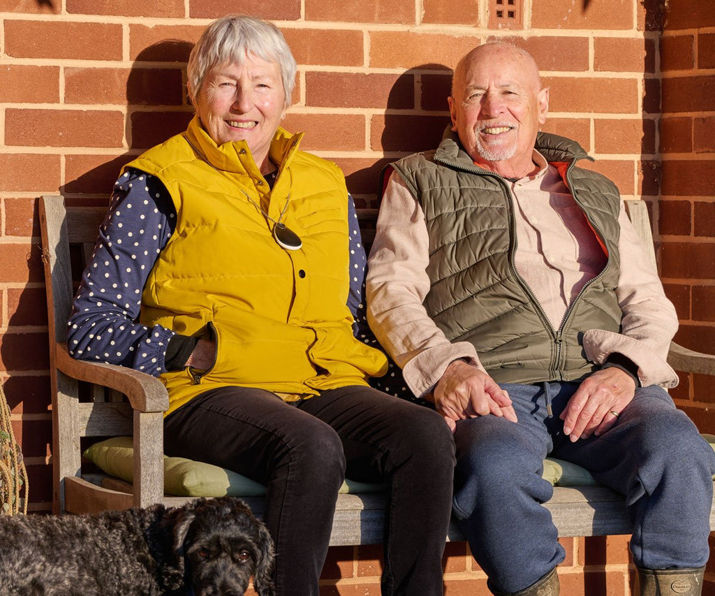 Two elderly people sitting on a bench in front of a brick wall, wearing personal alarms