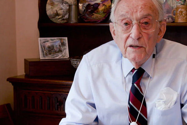 Older man wearing glasses and a white shirt with a tie, sitting in an indoor setting.