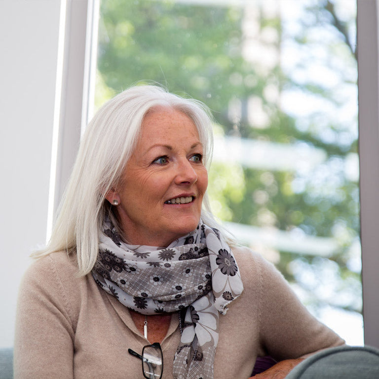 Older woman wearing a patterned scarf sitting indoors with a blurred outdoor background