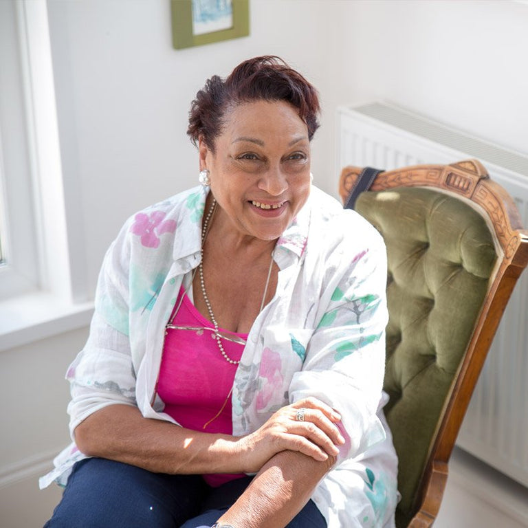 Woman sitting in a chair wearing a white floral shirt over a pink top, smiling.