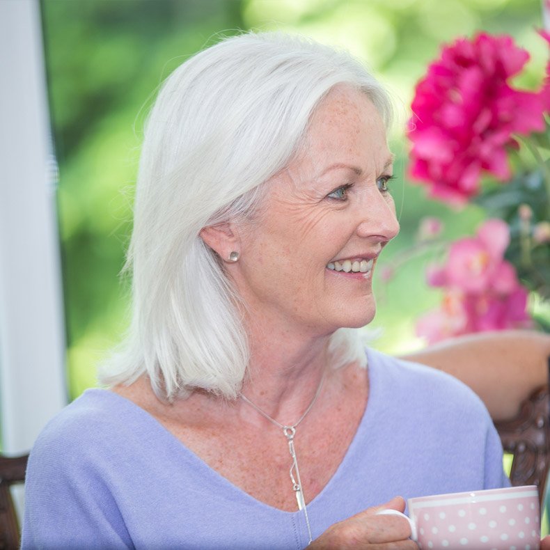 Woman with white hair holding a cup outdoors with flowers in the background